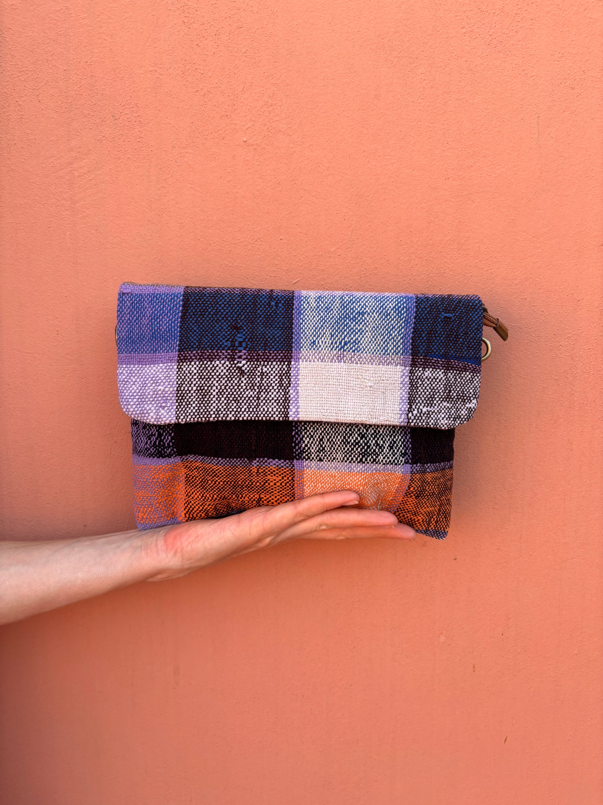 Eine Hand hält eine rechteckige Vintage Clutch von LEY! mit einem blauen, lila, weißen und orangefarbenen Karomuster aus upgecycelter Wolle vor einer einfarbigen pfirsichfarbenen Wand.