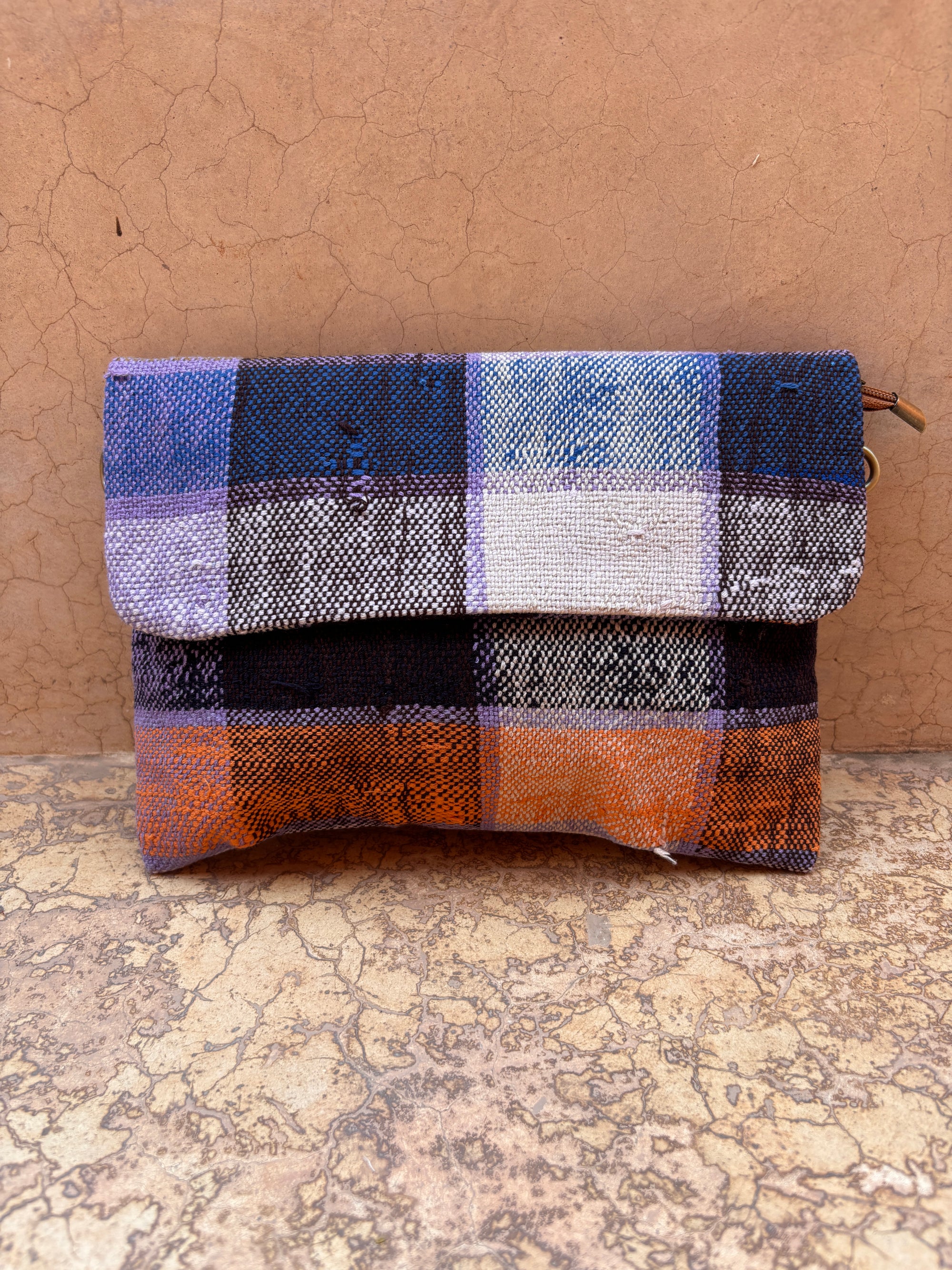 LEY! Vintage Clutch - eine handgefertigte kleine Stofftasche mit Klappverschluss, die ein blau, orange, lila und weiß kariertes Muster aufweist. Ideal als stilvolles Accessoire, das vor einem erdfarbenen, strukturierten Hintergrund gezeigt wird.