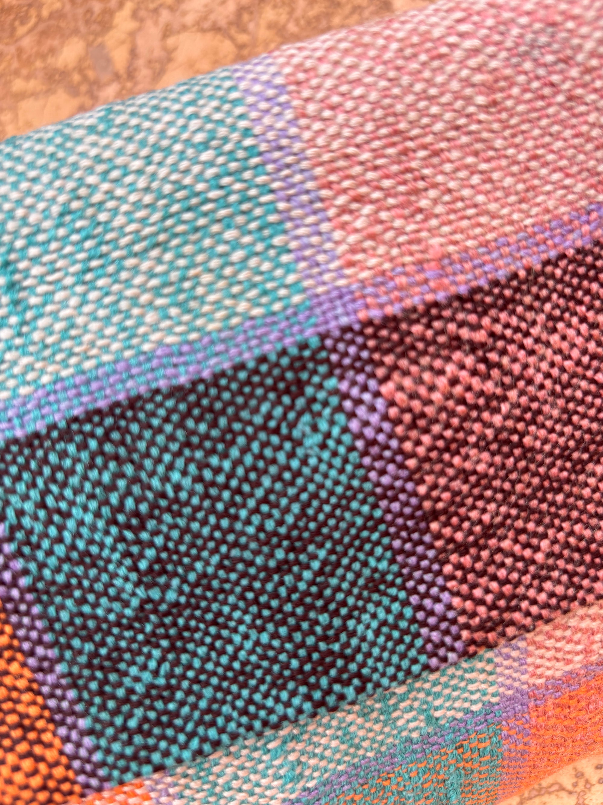 Eine Nahaufnahme eines farbenfrohen karierten Stoffes mit blaugrünen, lila, orangefarbenen und rosafarbenen Quadraten hebt die gewebte Textur hervor - ideal für die Herstellung der LEY! Vintage Clutch. Die leuchtenden Wollstoffe heben sich von einem sanft verschwommenen Hintergrund ab.