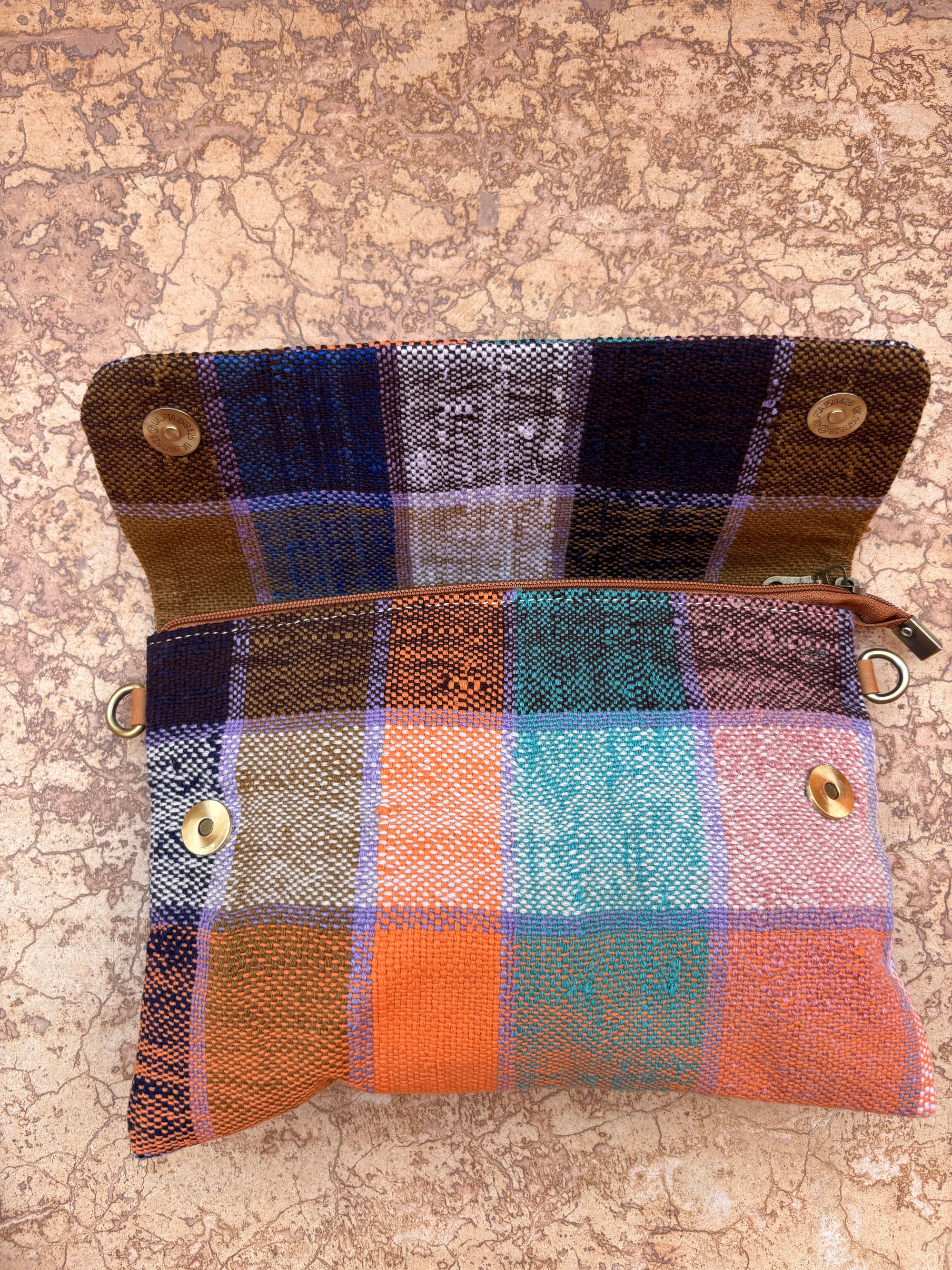 Die LEY! Vintage Clutch ist eine bunte, gewebte Tasche in karierten Orange-, Blau-, Türkis-, Lila- und Brauntönen. Diese handgefertigte Tasche verfügt über Metalldruckknöpfe, ein Reißverschlussfach und liegt offen auf einer strukturierten Oberfläche.
