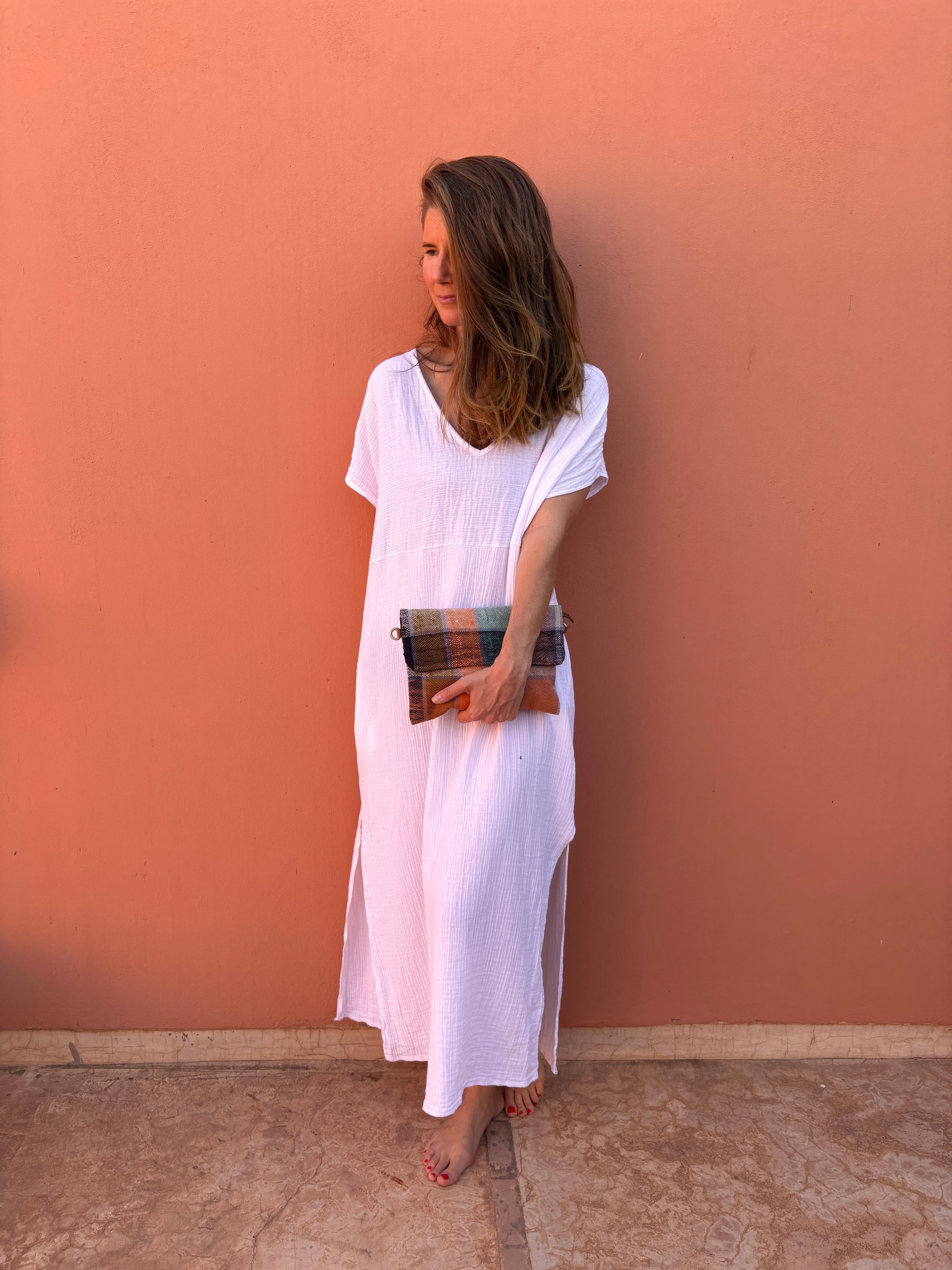 Eine Frau steht barfuß vor einer pfirsichfarbenen Wand, ihr Haar ist offen und sie strahlt mühelosen Stil aus, während sie die LEY! Vintage Clutch hält, während sie ein weites weißes Kleid trägt.