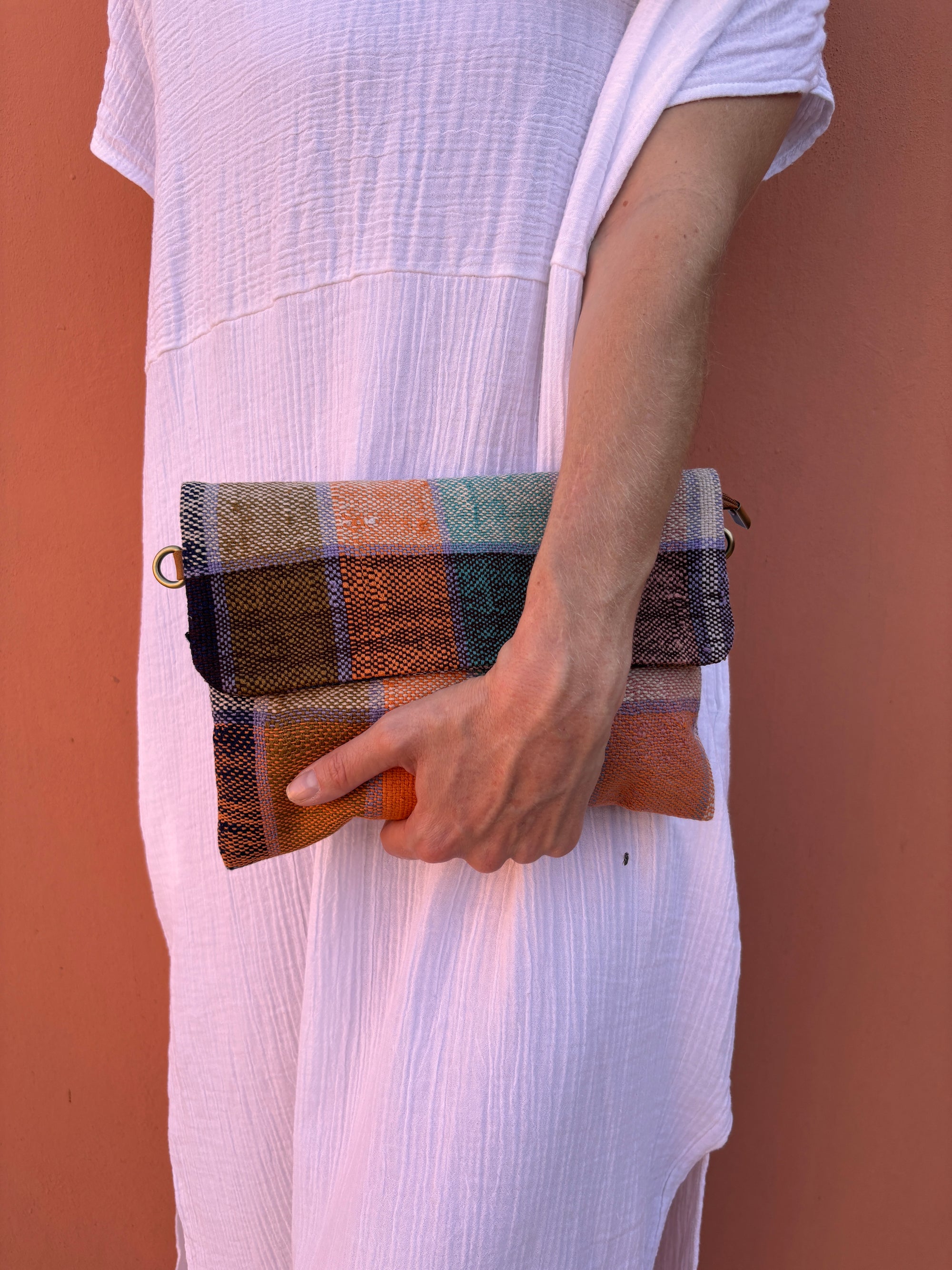 Eine Person in einem weißen, strukturierten Kleid hält die bunte LEY! Vintage Clutch gegen eine korallenrote Wand, den Arm entspannt an der Seite.