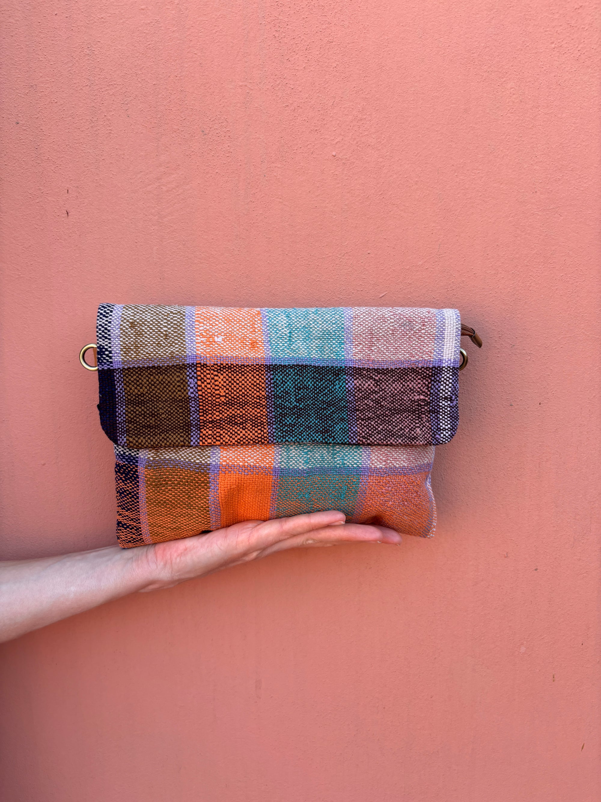 Eine Hand hält die Vintage Clutch von LEY! - eine bunte, rechteckige Tasche mit einem orangefarbenen, blauen, grünen, braunen und beigen Karomuster - vor einer pfirsichfarbenen Wand.