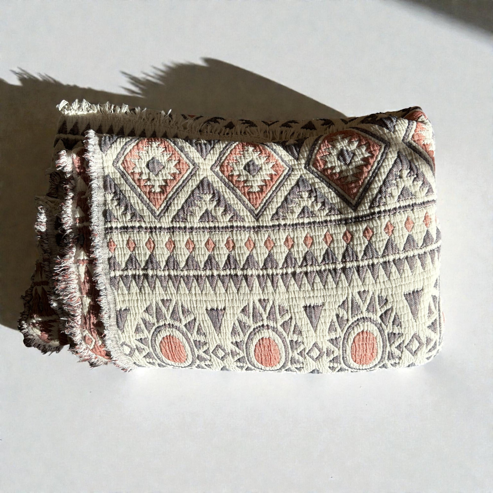 Die CALMA Musselin Decke von LEY! in Rot-Grau-Weiß (2,30 x 2,60 m) zeigt ein geometrisches Boho-Design mit Rauten und Ovalen sowie ausgefransten Kanten, das auf einer weißen Oberfläche mit weichem Schatten ruht.
