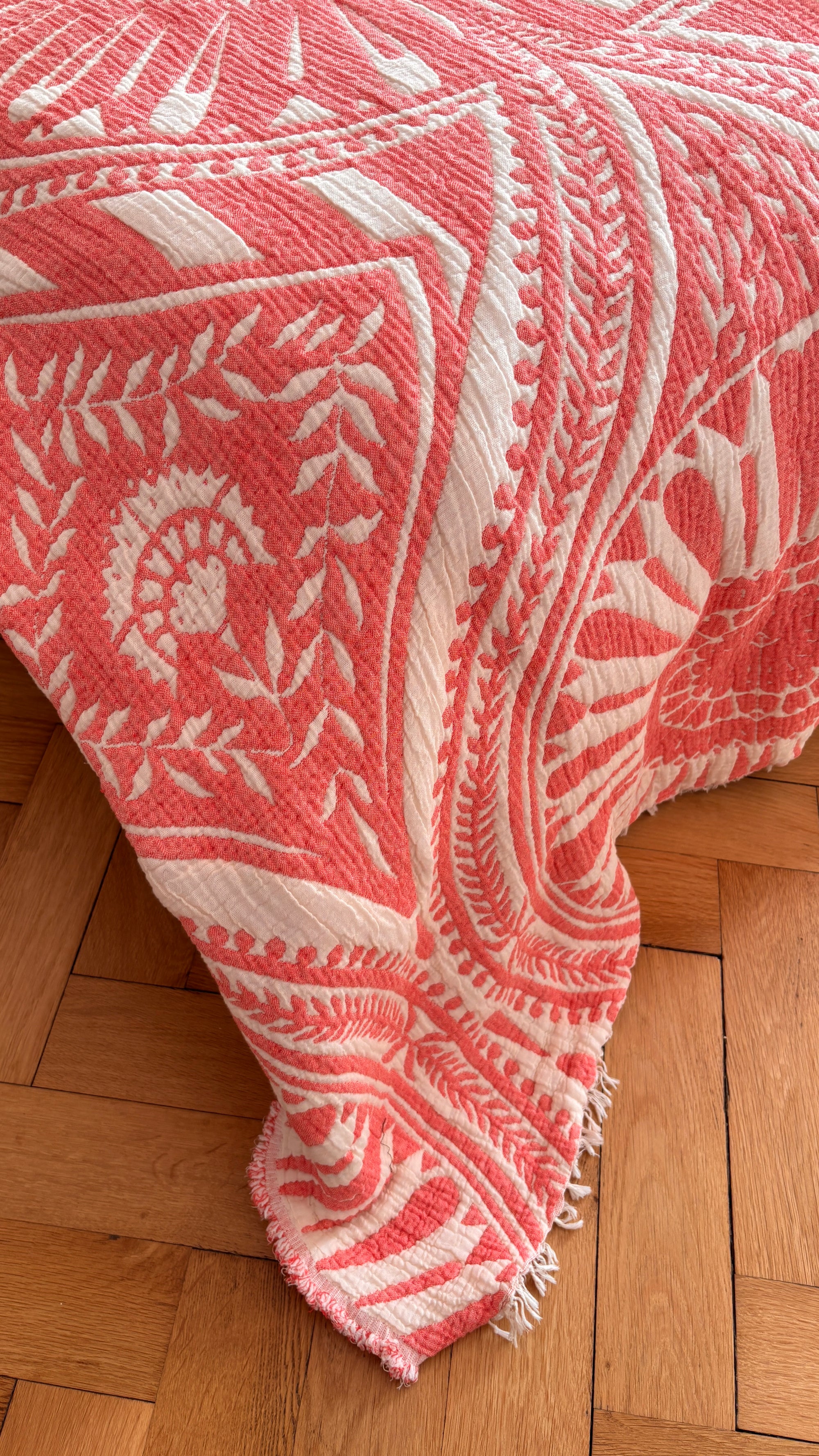 Die CALMA Musselin Decke - Koralle von LEY! (2,20 x 2,30 m) zeigt ein rosa-weißes Boho-Muster. Die atmungsaktive, strukturierte und gefranste Decke lässt sich über die Bettkante drapieren und liegt auf einem hellen Holzparkettboden.