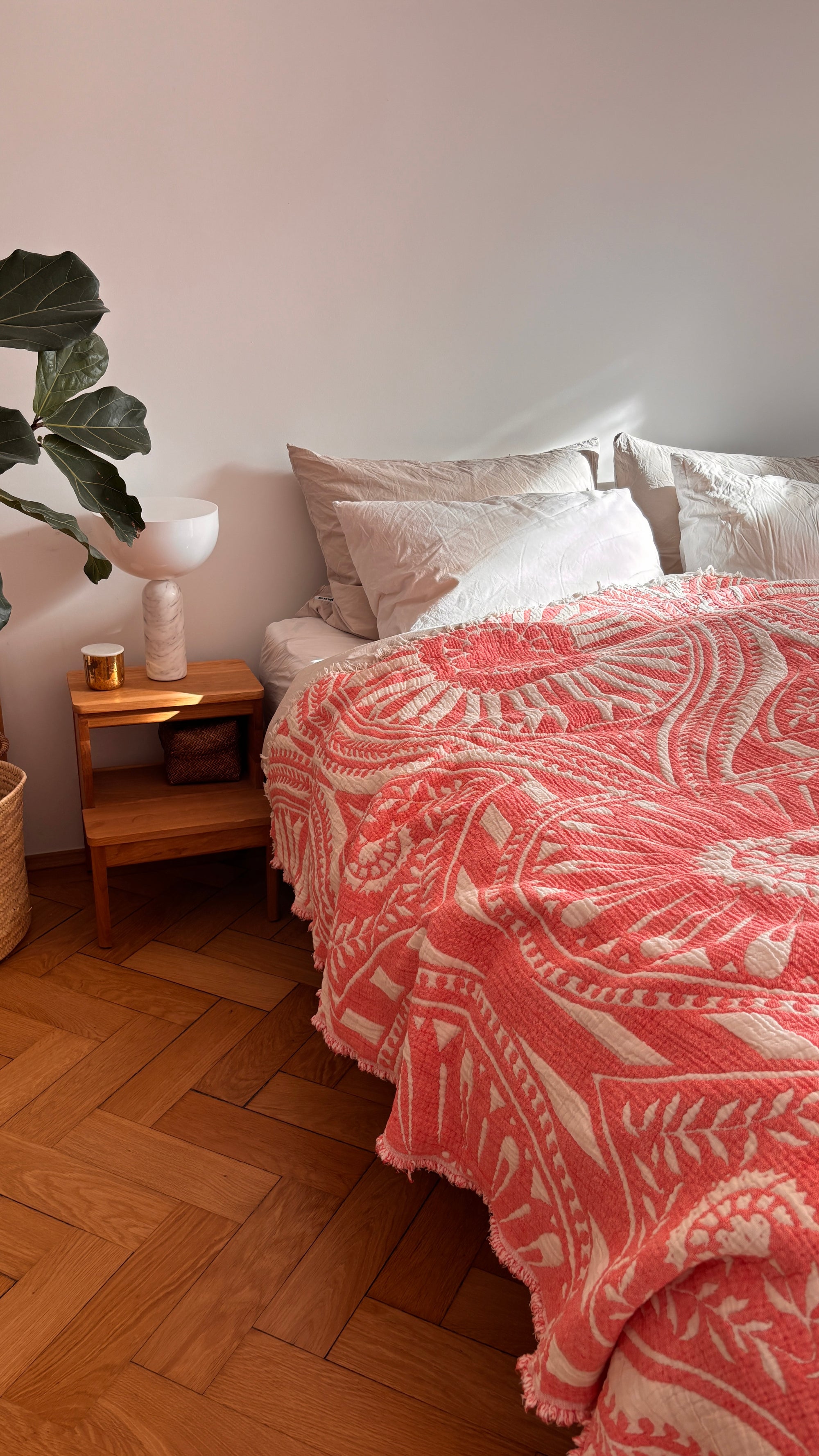 Eine gemütliche Schlafecke mit einer LEY! CALMA Musselin Decke in Koralle (2,20 x 2,30 m), weißen Kissen, einem hölzernen Nachttisch, einer Lampe, einer Kerze, einem Glas und einer grünen Blattpflanze auf dem Parkettboden - ideal für Liebhaber des Boho-Designs.