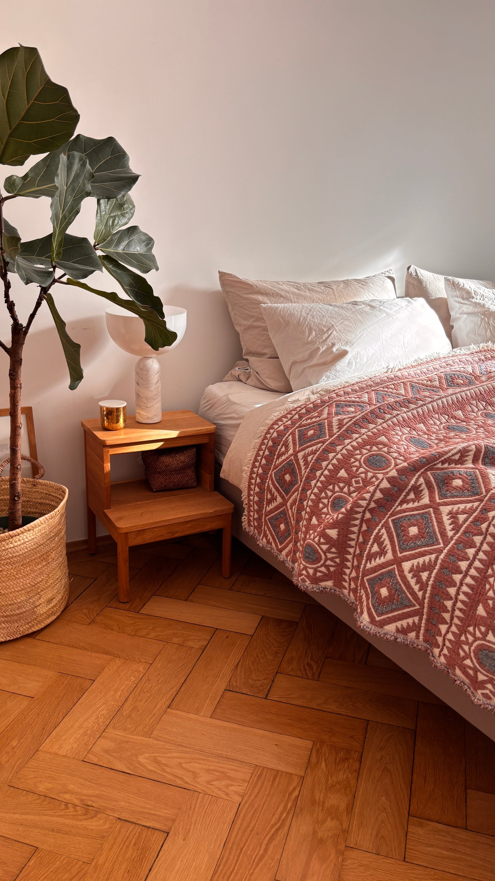 Ein gemütliches Schlafzimmer im Boho-Design - mit einem Bett, das mit der LEY! CALMA Musselin Decke in Rot-Grau-Weiß (2,30 x 2,60 m), weißen Kissen, einem hölzernen Nachttisch mit Dekostücken, einer getopften Feigenpflanze und einem Holzboden mit Fischgrätenmuster.