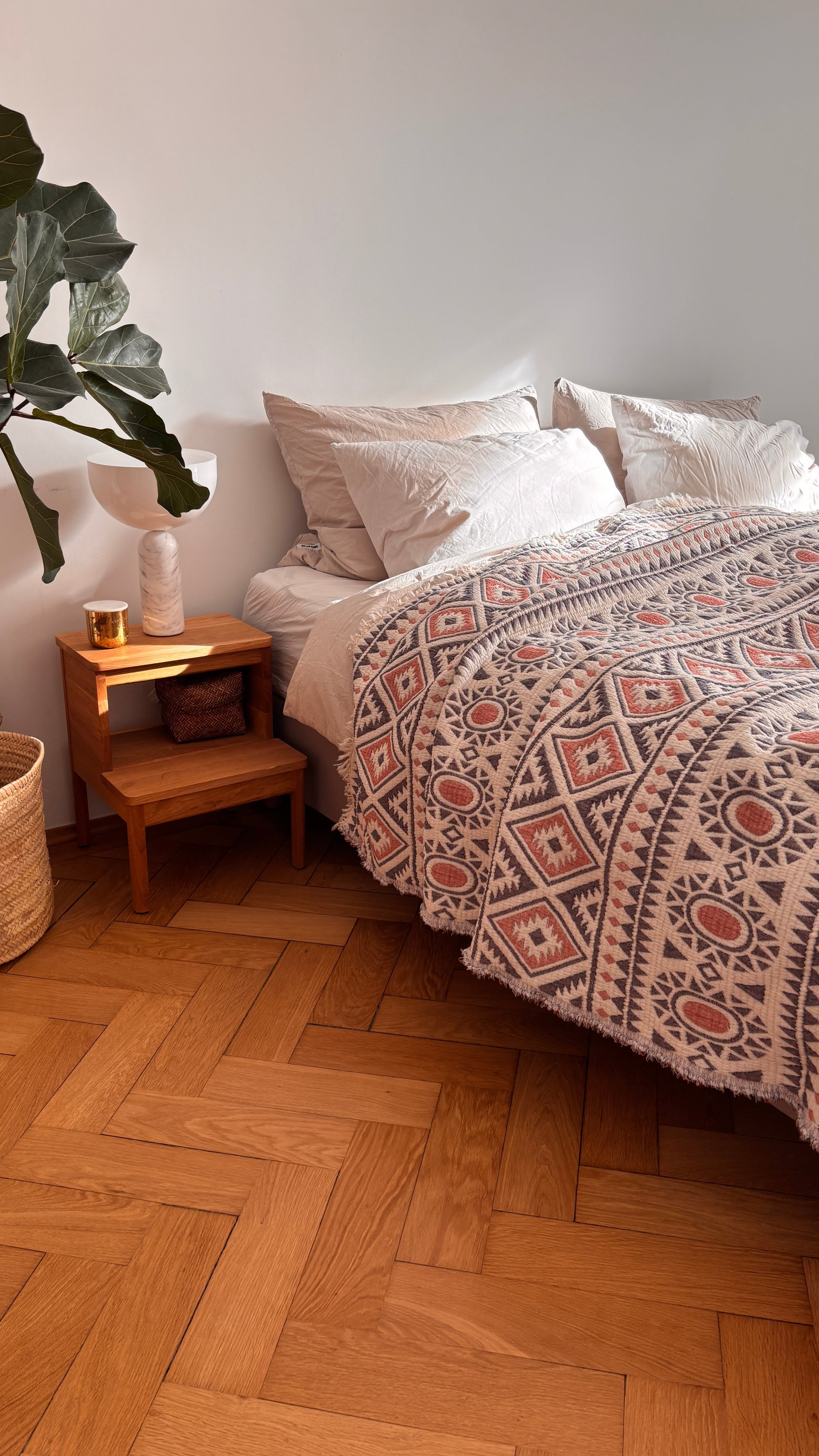 Ein gemütliches Schlafzimmer mit der LEY! CALMA Musselin Decke in Rot-Grau-Weiß (2,30 x 2,60 m), weißen Kissen, einem hölzernen Nachttisch mit Kerze und Vase, einer Topfpflanze und einem geflochtenen Korb auf einem Holzboden.