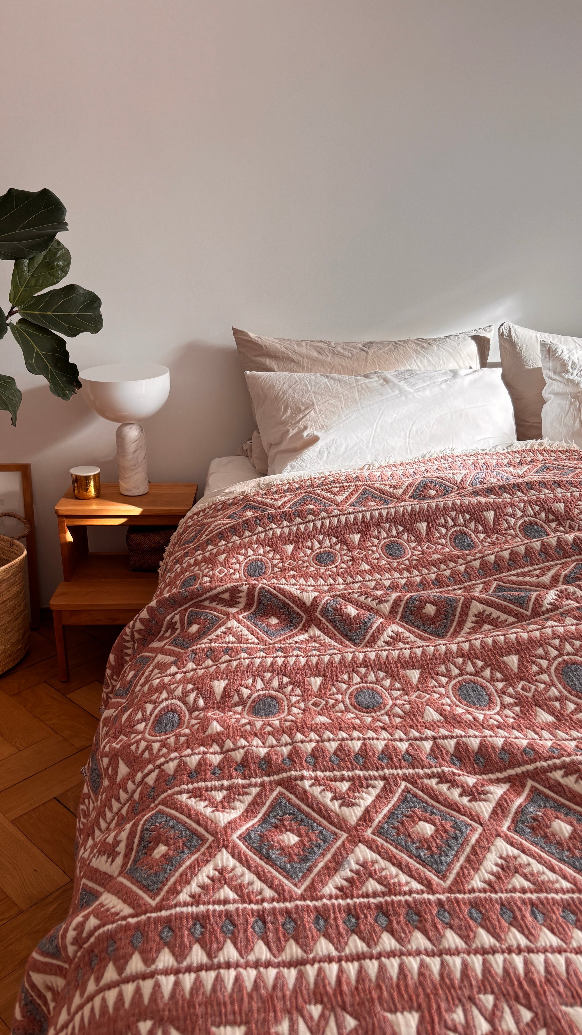 Ein gemütliches Bett mit weißen Kissen ist mit der LEY! CALMA Musselin Decke in Rot-Grau-Weiß (2,30 x 2,60 m) und sitzt neben einem hölzernen Nachttisch mit Pflanze, Büchern, Kerze und Lampe vor einer hellen Wand.