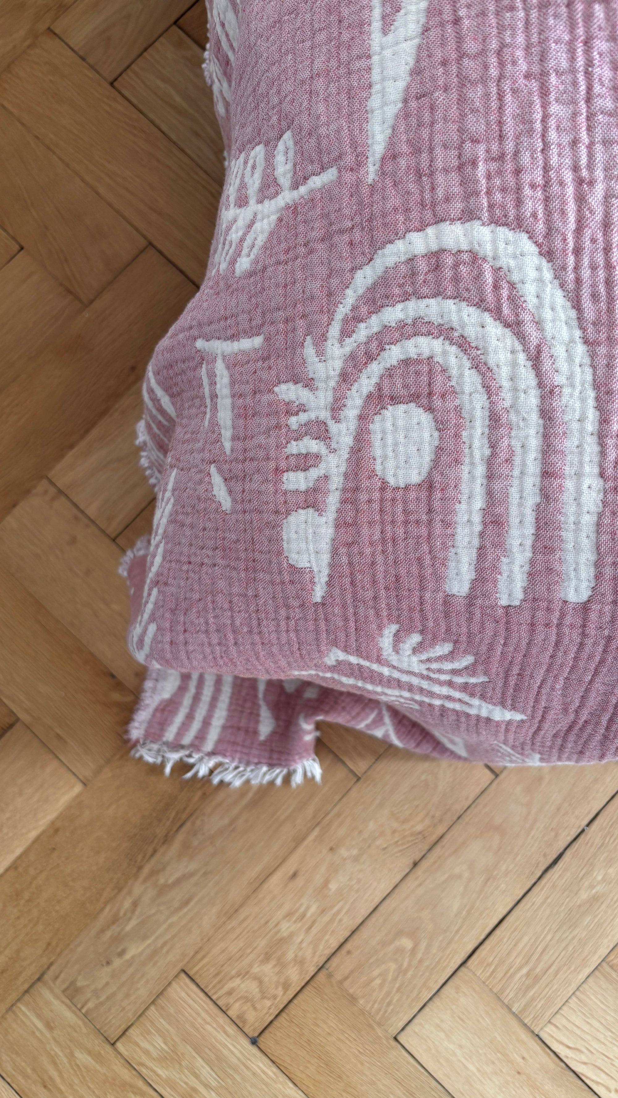 Die CALMA Musselin Decke von LEY! in rotbraun-weiß (1,60 x 2,30 m) mit ausgefransten Kanten liegt teilweise gefaltet auf dem Fischgrät-Holzboden und verleiht der gemütlichen Szene einen luftigen, atmungsaktiven Touch.