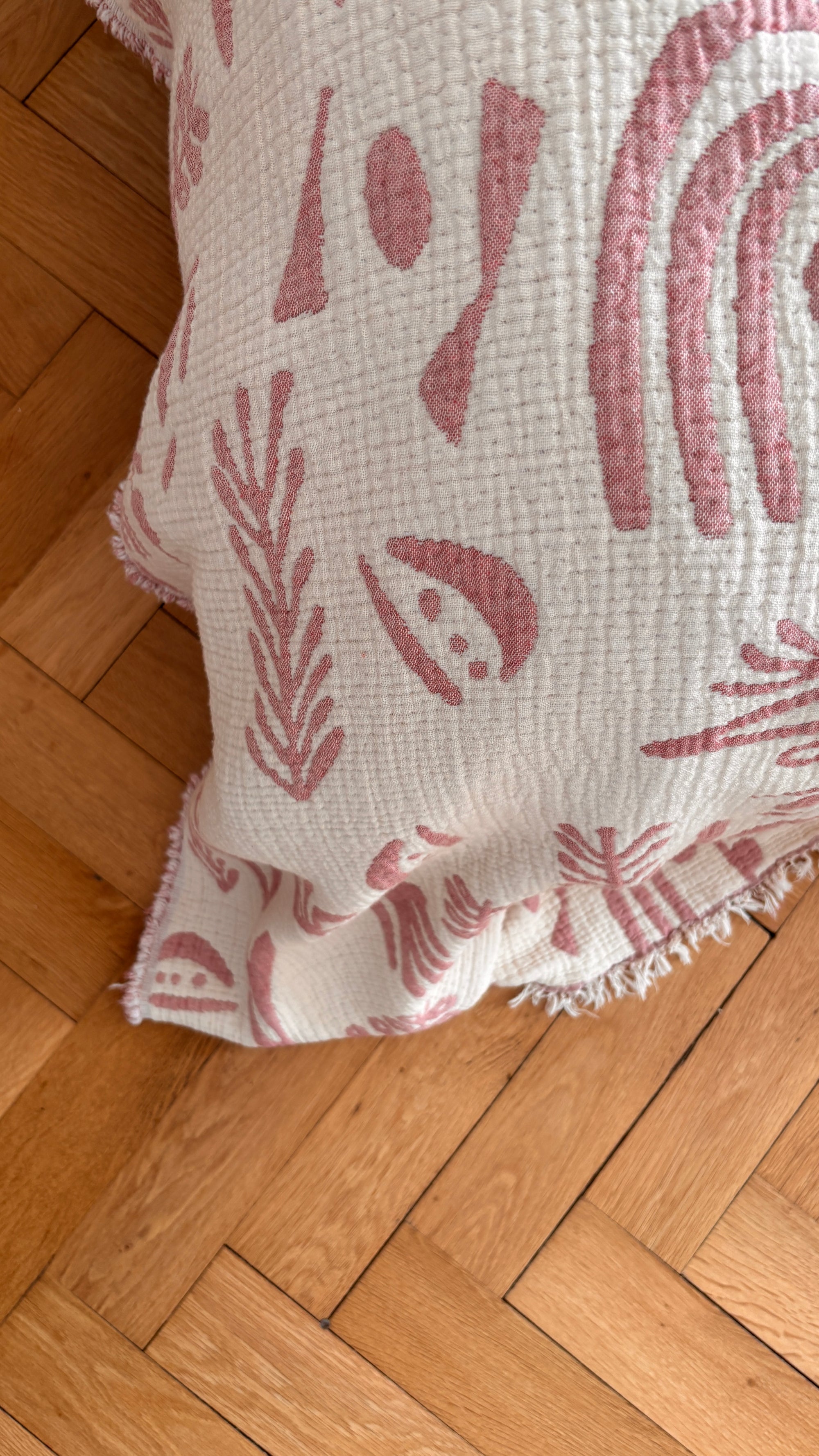 Die CALMA Musselin Decke von LEY! in rotbraun-weiß (1,60 x 2,30 m) mit abstrakten Motiven und Fransenkanten bringt gemütliches Boho-Design auf einen hellen Holz-Fischgrätboden.