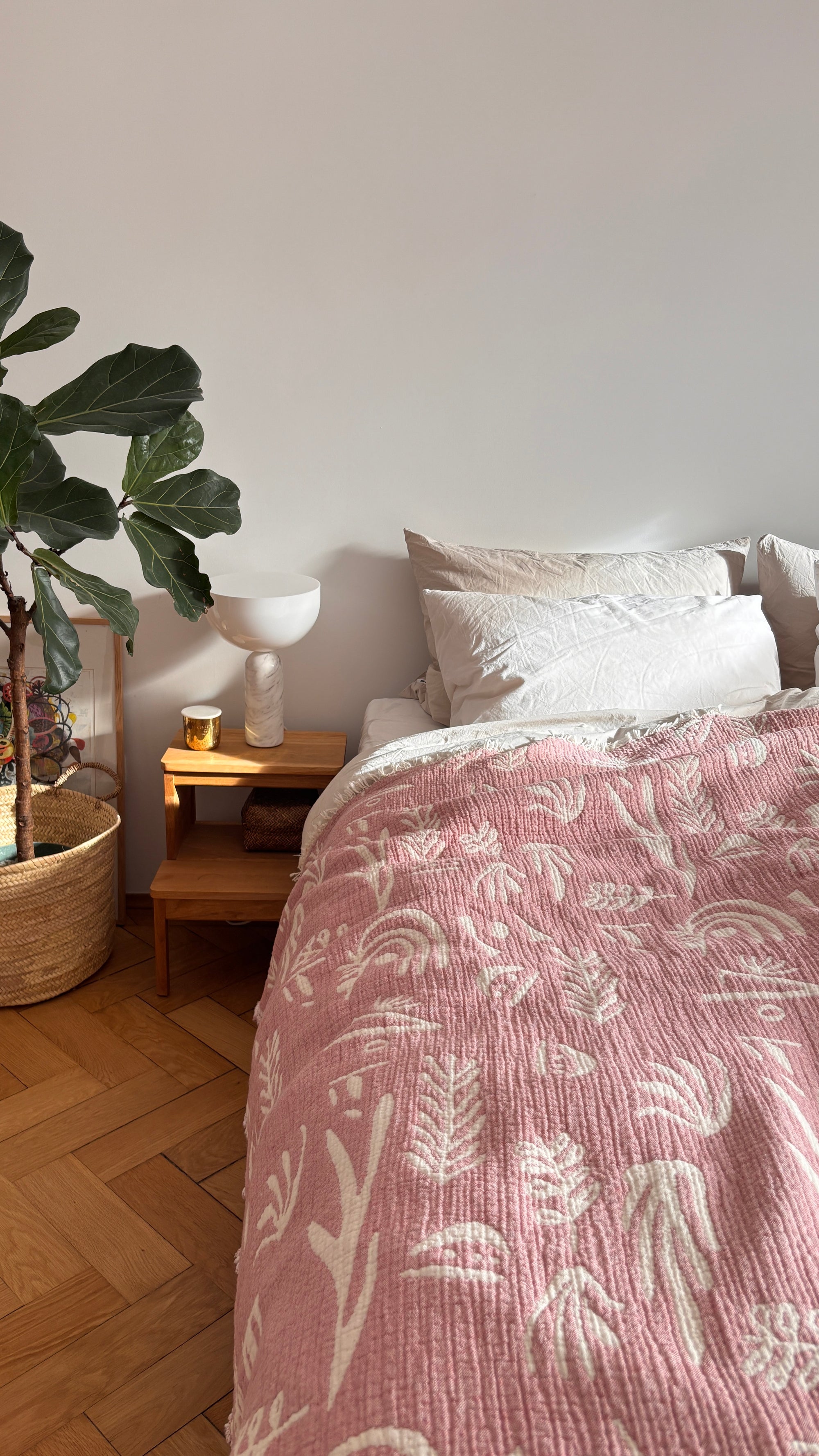 Ein gemütliches Schlafzimmer mit einem Bett, das mit der LEY! CALMA Musselin Decke in Rotbraun-Weiß (1,60 x 2,30 m), mit weißen botanischen Mustern, weißen Kissen, einem hölzernen Nachttisch mit Kerze und Dekoration und einer großen Topfpflanze auf Holzboden.