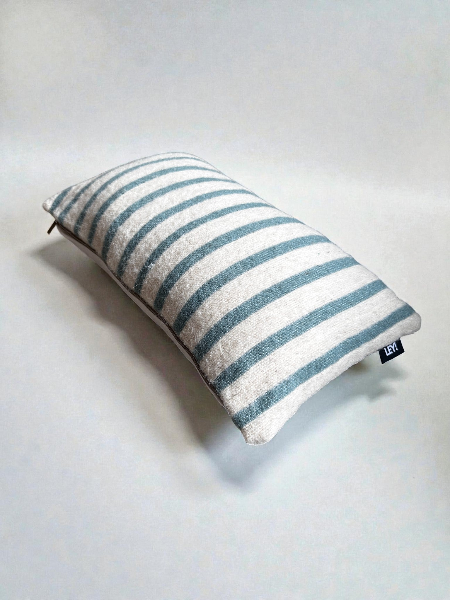 Ein rechteckiges LEY! Kissen - WINTER EDITION - Lagune - 30x50 mit hellblauen und weißen Streifen ruht auf einem schlichten weißen Hintergrund. Ein kleines schwarzes Etikett mit weißem Text ist an einer Ecke dieses Kissens angenäht.