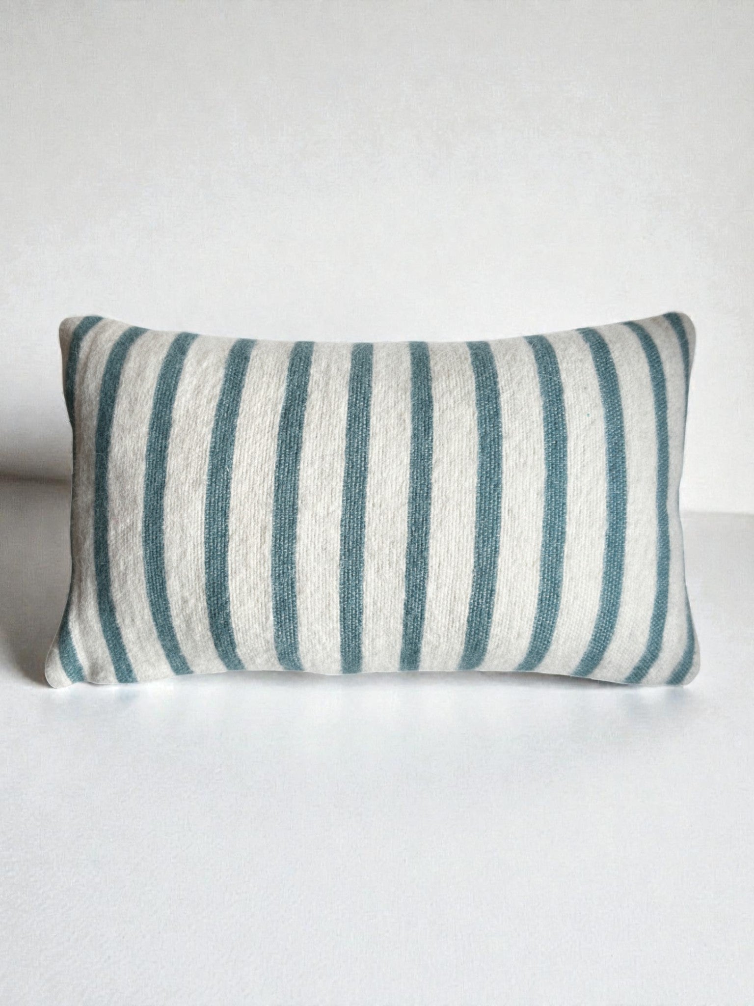 Ein rechteckiges LEY! Kissen - WINTER EDITION - Lagune - 30x50 mit blauen und weißen Längsstreifen, handgefertigt in Marrakesch, auf einer schlichten weißen Fläche vor weißem Hintergrund.