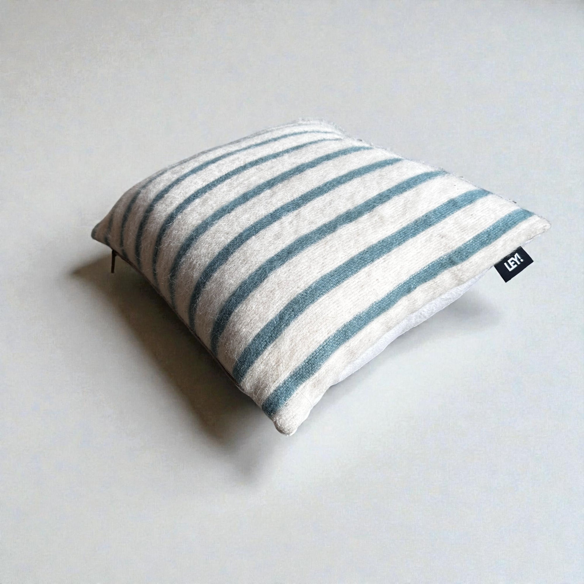 Ein quadratisches Kissen - WINTER EDITION - Lagune - 40x40 von LEY! mit weißen und blauen Querstreifen ruht auf einer hellgrauen Oberfläche, auf der ein kleines schwarzes Stoffschild mit weißem Text angebracht ist.