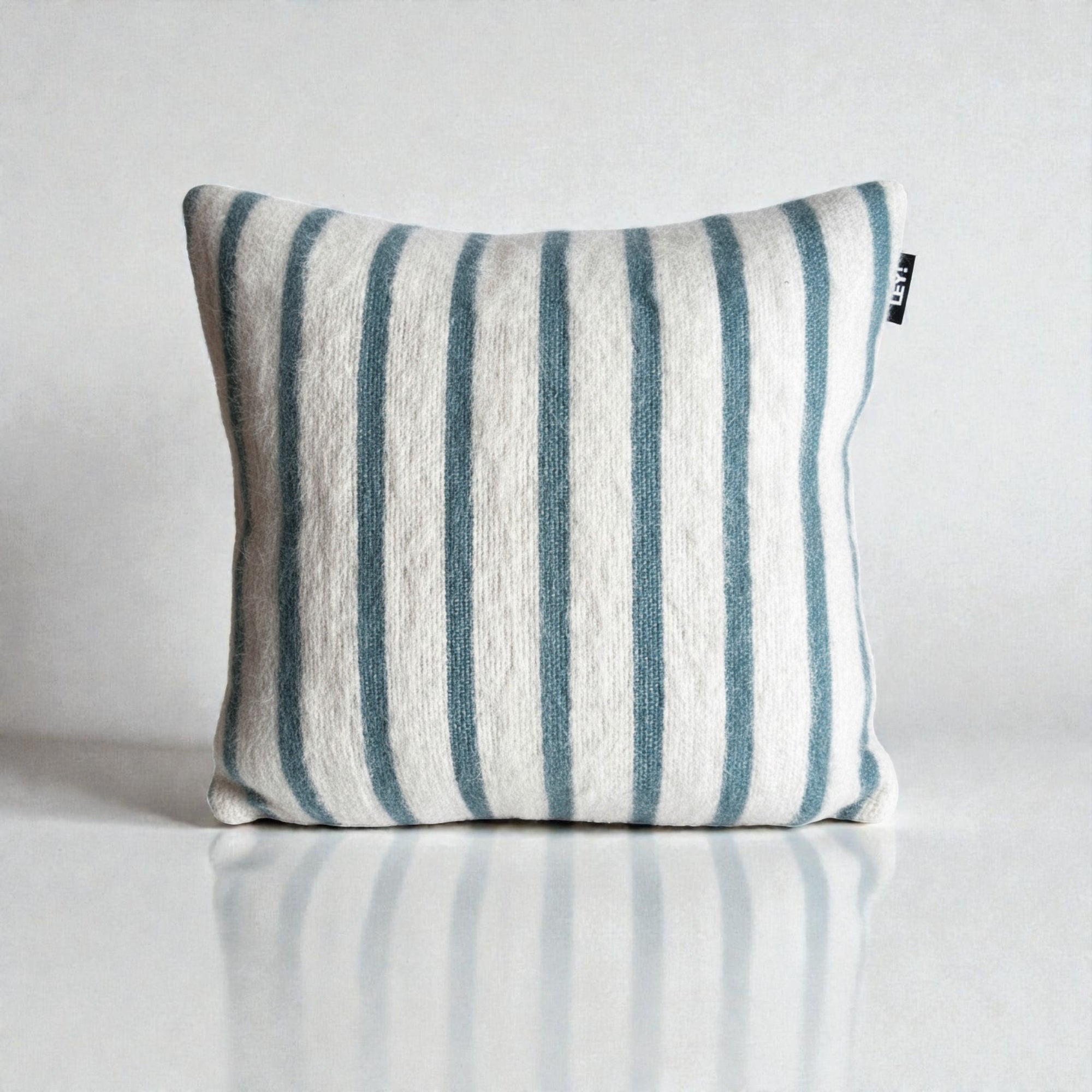 Das Kissen - WINTER EDITION - Lagune - 40x40 von LEY! hat eine quadratische Form, einen weißen Hintergrund mit vertikalen blaugrauen Streifen, einen weichen Schurwolle-Stoff, ein kleines schwarzes Etikett auf der rechten Seite und zeigt die Handwerkskunst von Handarbeit Marrakesch.