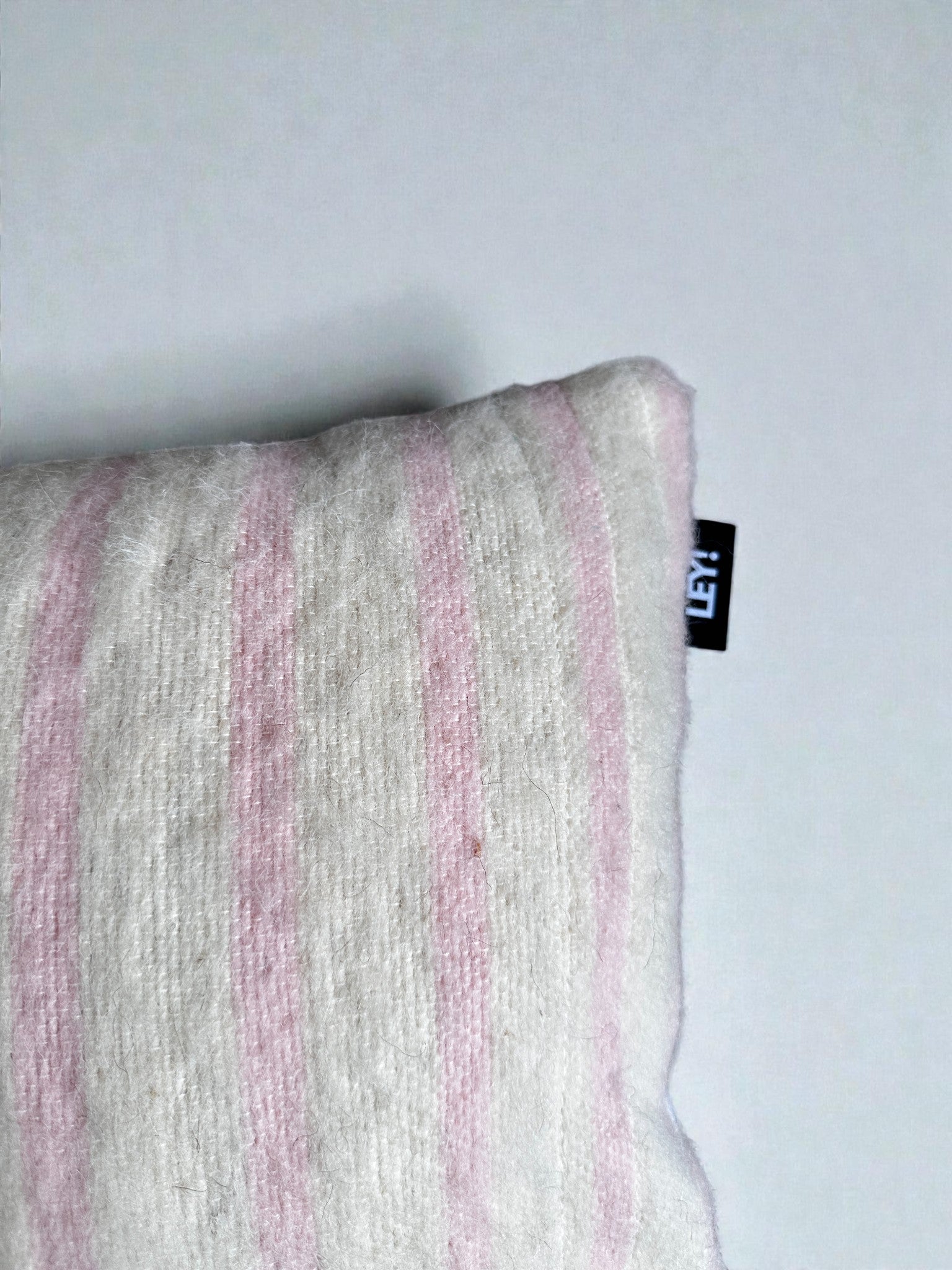 Eine Nahaufnahme des LEY! Kissen - WINTER EDITION - Puder 30 x 50 zeigt weiche, hellrosa und weiße Streifen auf strukturiertem Stoff mit einem kleinen schwarzen Anhänger am rechten Rand vor einem hellen Hintergrund.