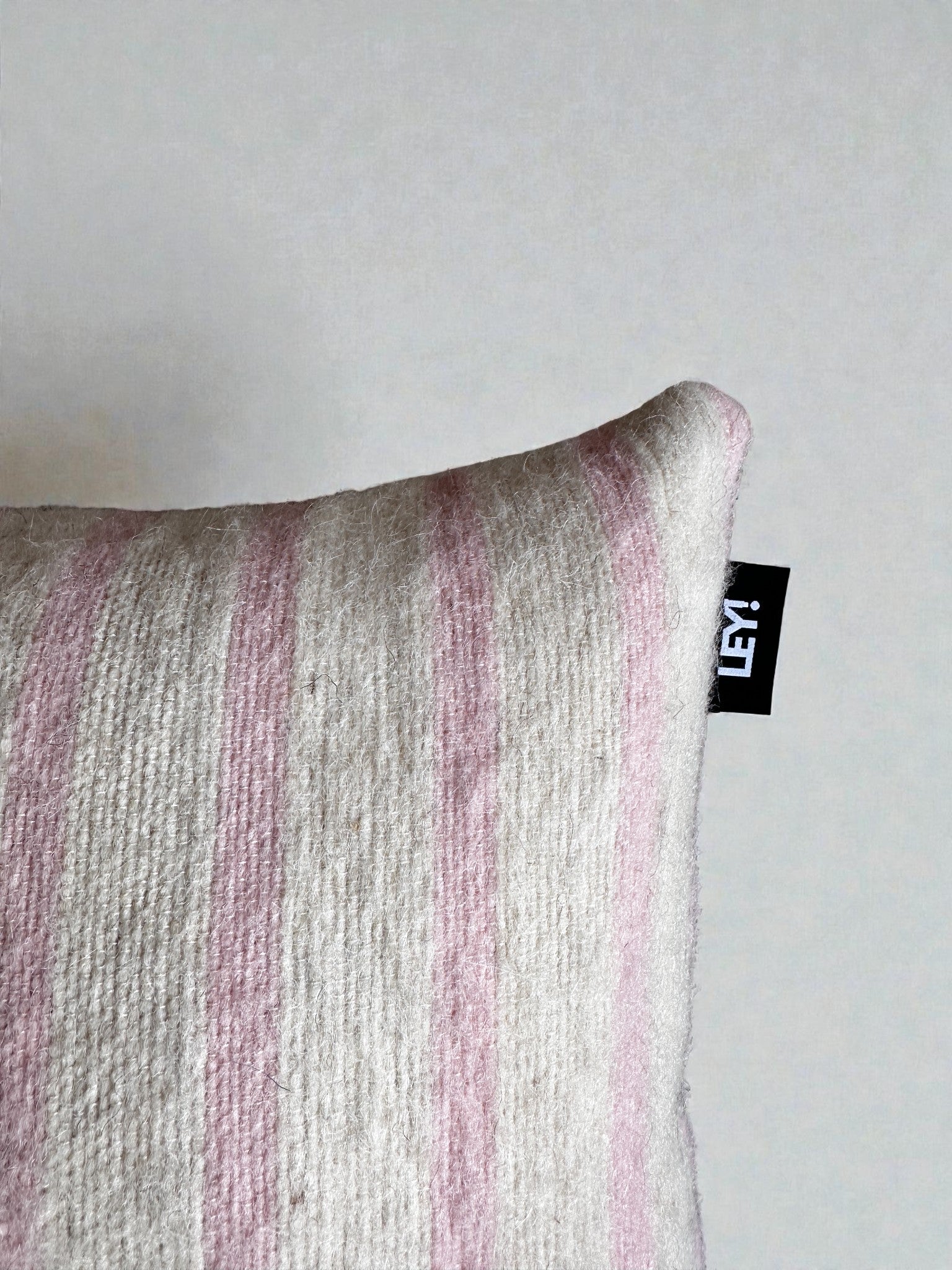 Eine Nahaufnahme des Kissens - WINTER EDITION - Puder- 40x40 von LEY! mit Schurwolle mit hellrosa und cremefarbenen Streifen, einem schwarzen "LEY!"-Schriftzug am oberen Rand und Details von Handarbeit Marrakesch vor einem hellen Hintergrund.