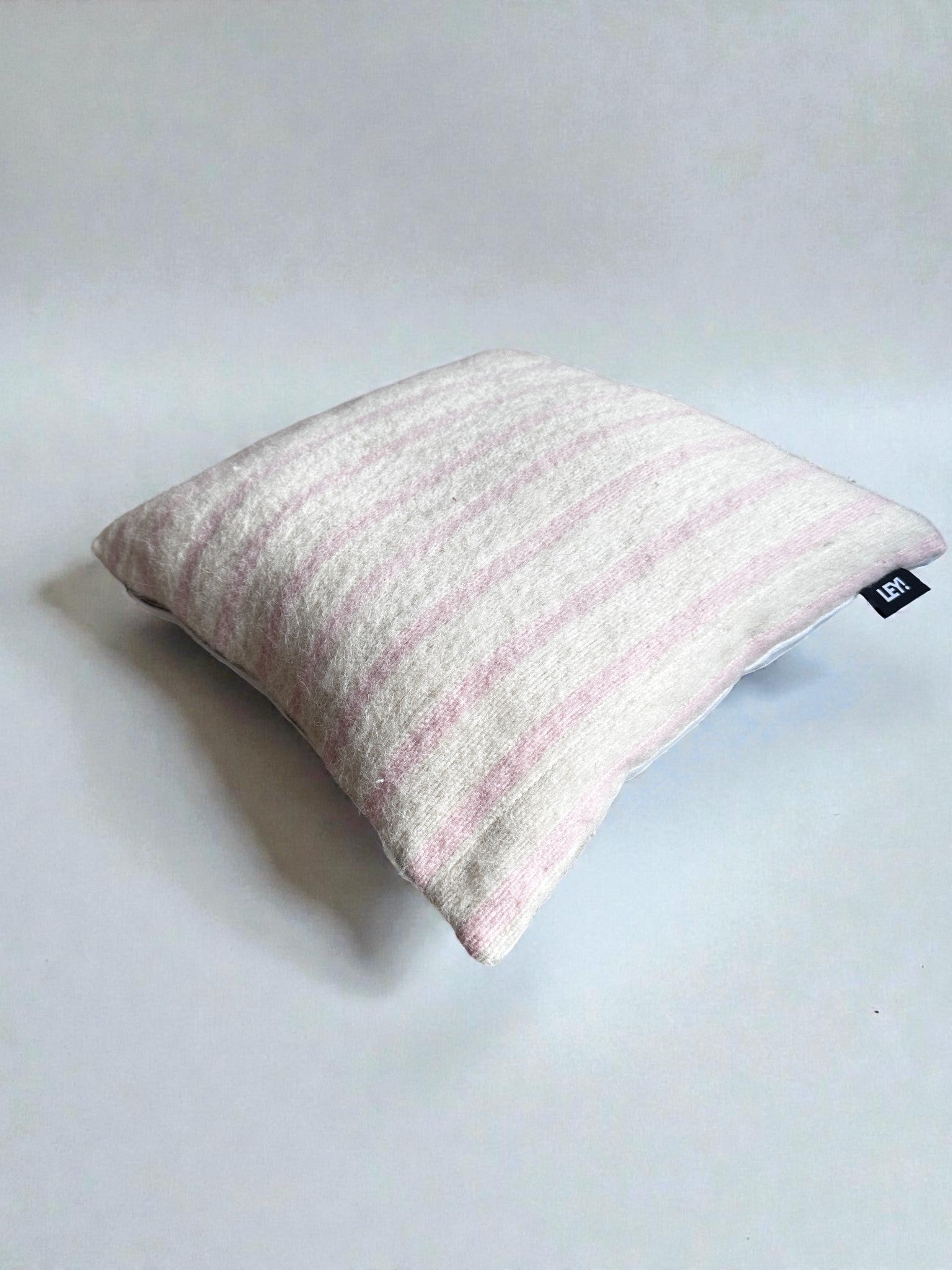 Ein quadratisches LEY! Kissen - WINTER EDITION - Puder- 40x40 mit hellrosa und weißen Streifen liegt auf einer unifarbenen weißen Fläche und hat ein kleines schwarzes Etikett an der Seite.