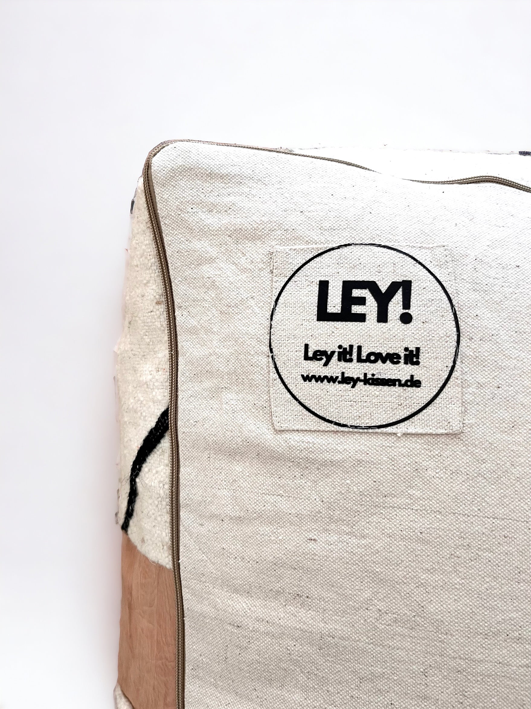Eine Nahaufnahme des LEY! Bodenkissen - Milu in beiger Schurwolle, mit braunem Rand, schwarzen Zierlinien und einem Aufnäher mit der Aufschrift "LEY! Ley it! Love it! www.ley-kissen.de". Handgefertigt mit viel Liebe zum Detail.