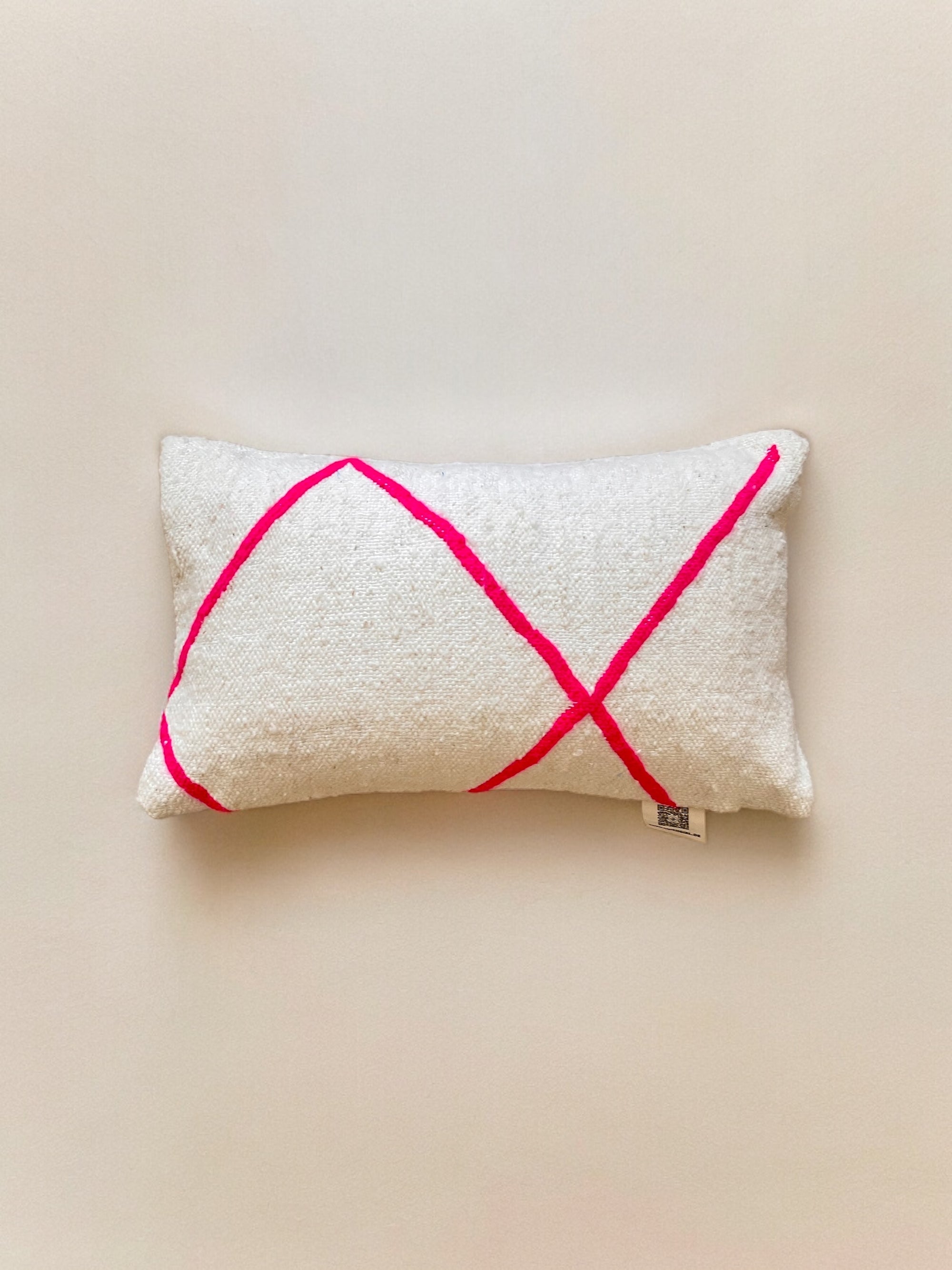 Das Kissen - Mina 40x60 von LEY! ist ein rechteckiger beiger Schurwolle-Kissenbezug mit rauer Struktur, handgemalten, sich kreuzenden roten Linien im X-Design und einem kleinen Stoffanhänger unten rechts. Handgemacht in Marrakesch.