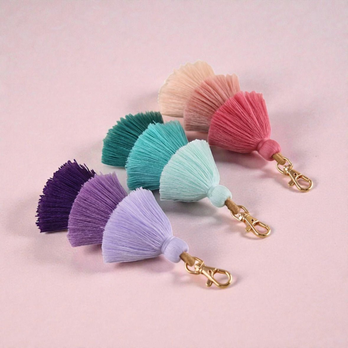 Vier Schlüsselanhänger QUASTE von LEY! in Lila, Blau, Türkis, Pink und Pfirsich liegen diagonal auf einem hellrosa Hintergrund. Mit goldener Hardware und fächerartigem Layer-Design - ein stylisches Accessoire und tolle Geschenkidee.