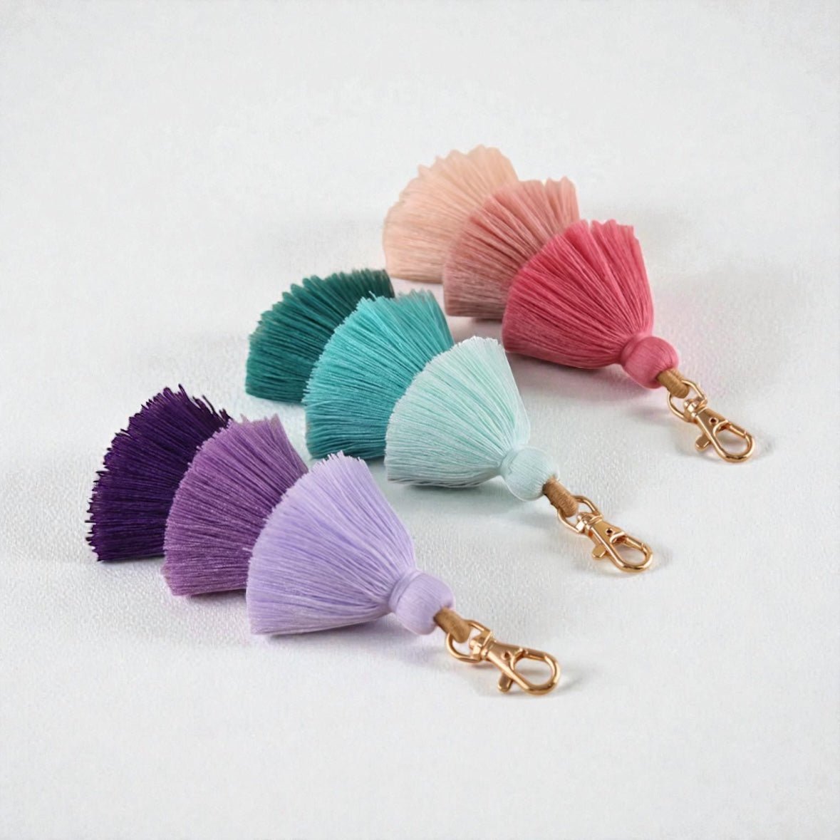 Vier bunte Schlüsselanhänger QUASTE von LEY! in Lila, Helllila, Türkis und Pink mit goldenen Verschlüssen sind diagonal auf weiß arrangiert - ein perfektes Accessoire und Geschenkidee.