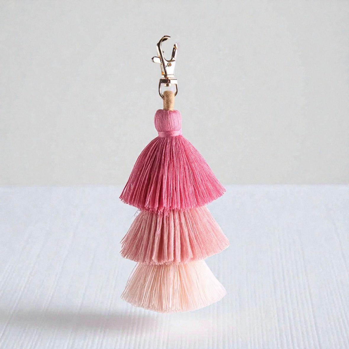 Der LEY! SCHLÜSSELANHÄNGER QUASTE zeigt vier Schichten rosa Fransen, von tiefen bis pastelligen Tönen, gekrönt von einer goldenen Schließe - eine auffällige Accessoire-Geschenkidee auf hellem Grund.