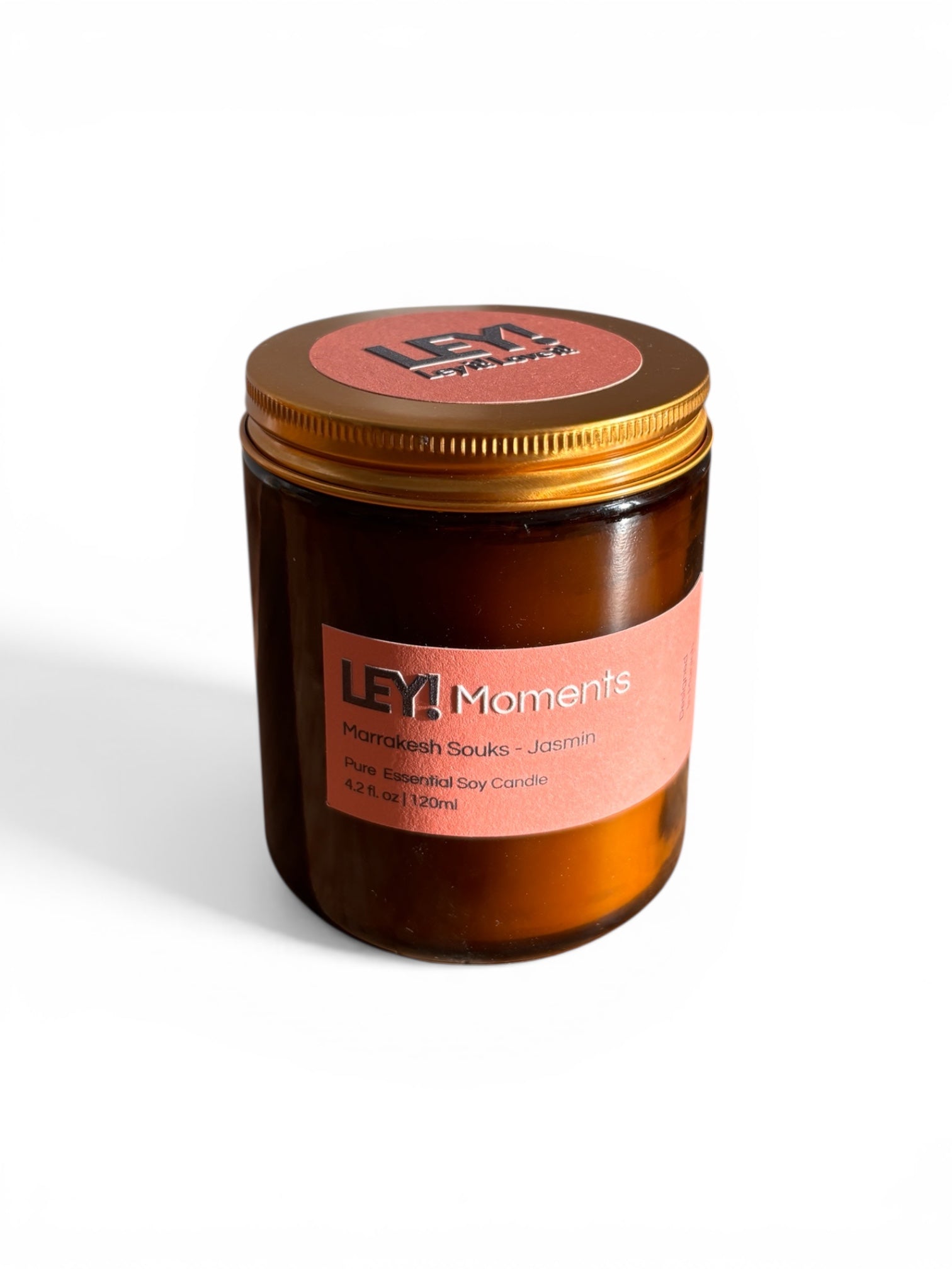 Eine braune Glaskerze mit einem goldenen Deckel und einem rosa Etikett mit der Aufschrift "LEY! Moments Marrakesh Souks - Jasmin Pure Essential Soy Candle, 4.2 fl. oz | 120ml" auf weißem Hintergrund.