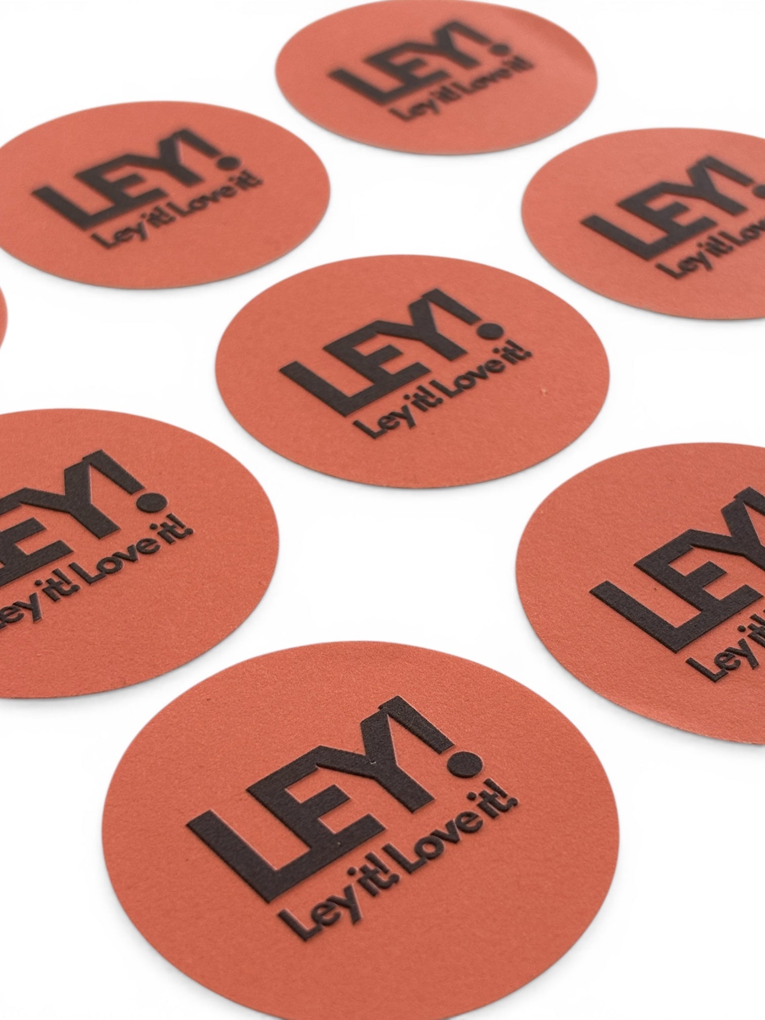 Mehrere runde, rötlich-orangefarbene LEY! STICKER mit fetten schwarzen "LEY! Ley it! Love it!"-Schriftzug sind auf einer weißen Fläche verteilt, perfekt für persönliches Branding wie einzigartige Logo-Aufkleber.