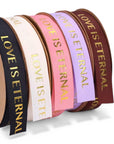 Fünf Rollen LEY! Bedrucktes Baumwollband in den Farben schwarz, weiß, rosa, lavendel und braun mit dem goldenen Text "LOVE IS ETERNAL". Ideal für Branding oder einzigartige Designs. Die Rollen sind in einer Reihe angeordnet, leicht überlappend.