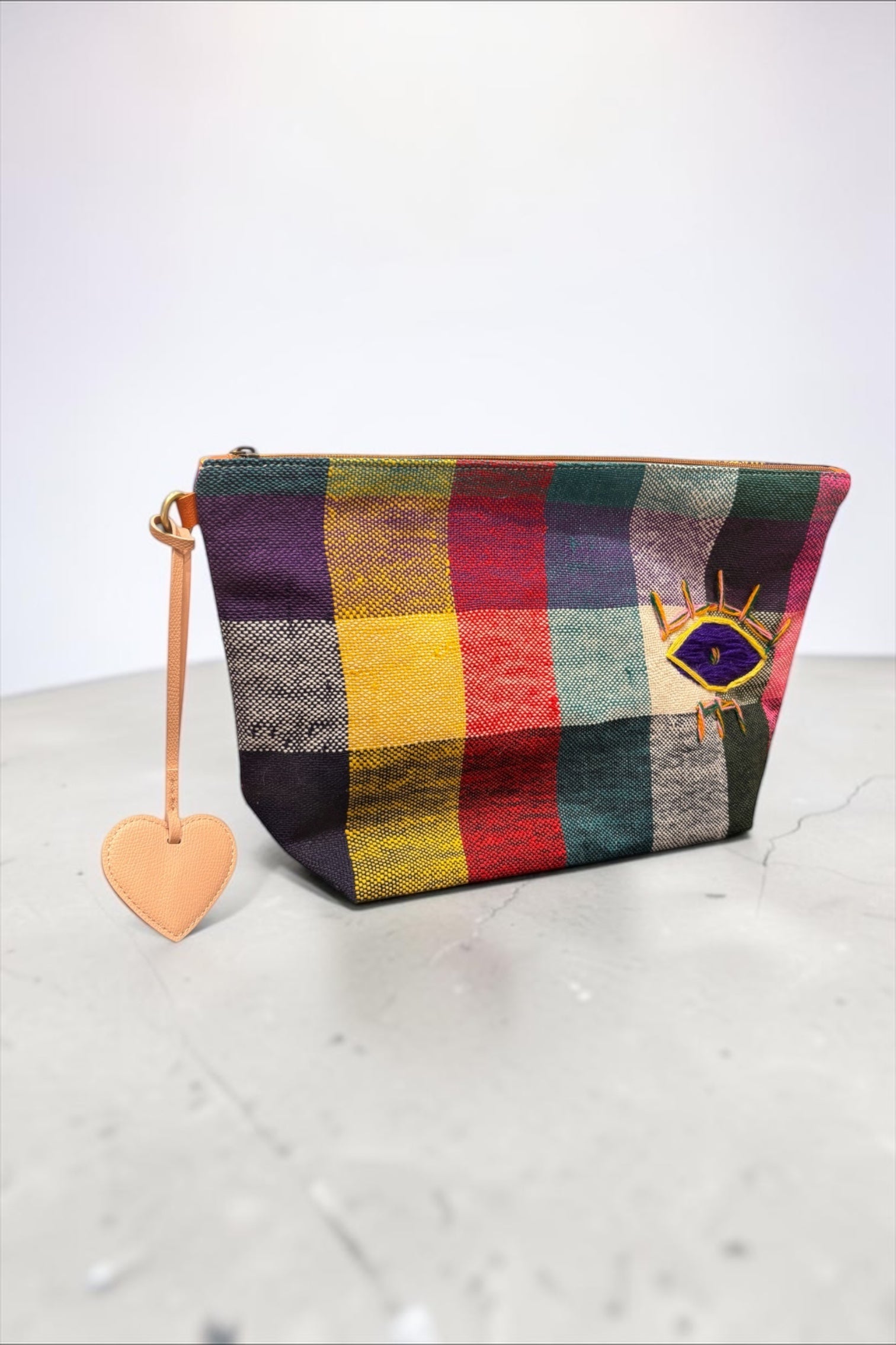 LEY! Taschenanhänger – Love Loop – Apricot: Ein karierter Beutel mit einem bestickten Augen- und Herzanhänger auf grauem Hintergrund.