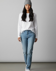 Eine Frau in einer beigen Unisex-Wollmütze „Saha“ von LEY!, einem weißen Langarmshirt und einer blauen Jeans steht vor einem einfarbigen Hintergrund.