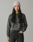 Frau mit langen schwarzen Haaren, die eine LEY!-Wollmütze „Saha“ in Pastellgrün, ein graues Sweatshirt und schwarze Jeans trägt.