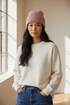 Eine Frau in einem cremefarbenen Pullover und einer LEY!-Wollmütze „Saha“ in Pink steht mit den Händen in den Taschen vor einem sonnendurchfluteten Fenster.