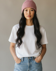 Eine Frau trägt eine LEY!-Wollmütze „Saha“ in Rosa, ein weißes T-Shirt und Jeans, die Hände in den Taschen, vor einem neutralen Hintergrund.