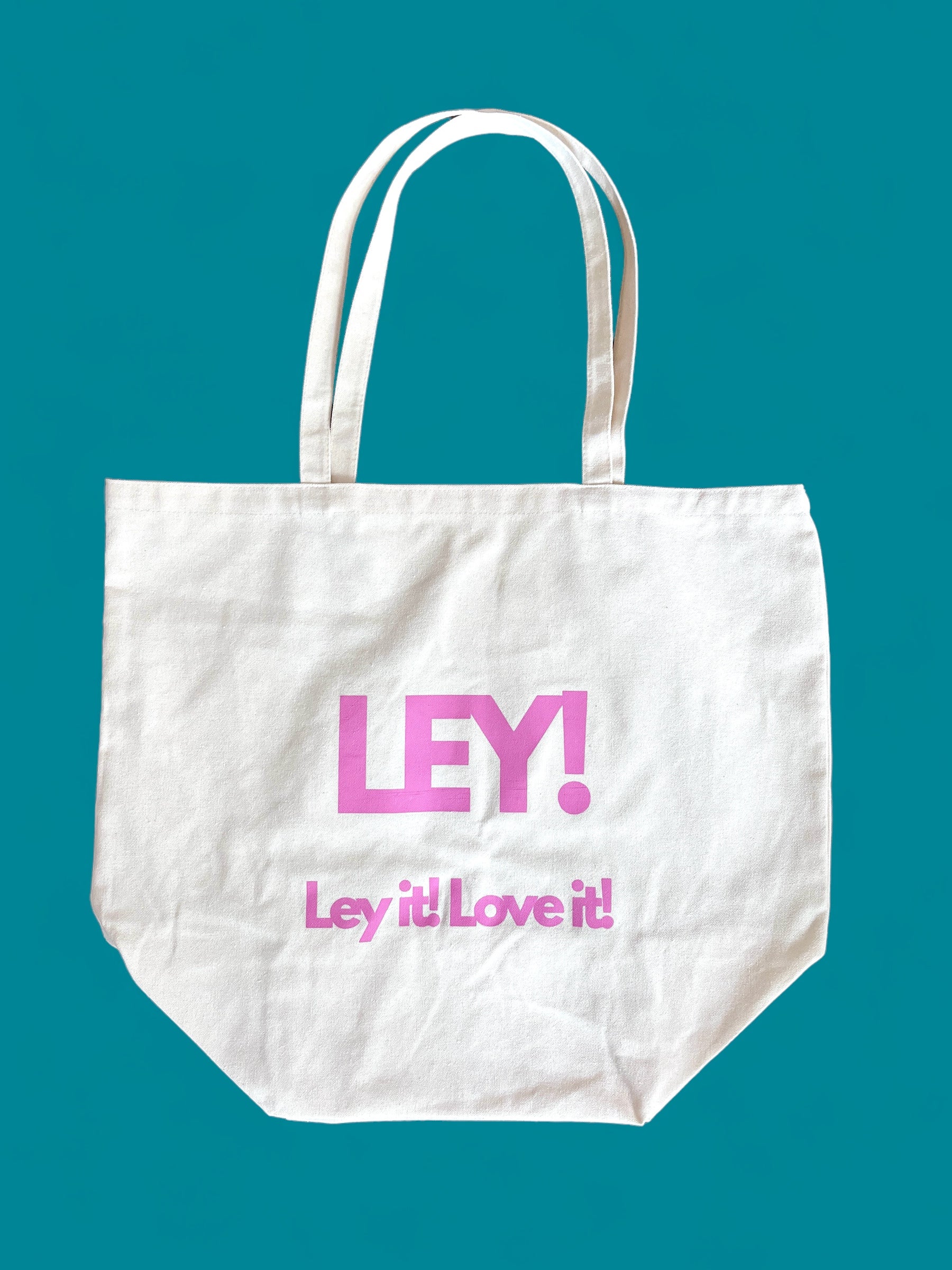 Eine weiße LEY! Tasche von LEY! mit zwei Henkeln steht auf einem tealfarbenen Hintergrund. Der fette rosa "LEY!"-Text erscheint über "Ley it! Love it!". Diese Baumwolltasche ist das perfekte Accessoire für jeden Tag.