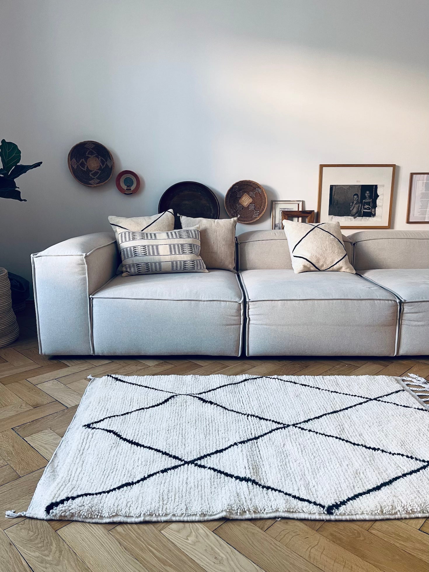 Ein modernes Wohnzimmer mit einem hellgrauen Sofa, gemusterten Kissen, runden Körben, gerahmter Kunst und dem BERBER TEPPICH - MAHIMA (1,50 x 2,50 m) von THE DANISH GIRL INTERIOR, einem weißen Wollteppich mit schwarzen geometrischen Linien auf Holzboden.