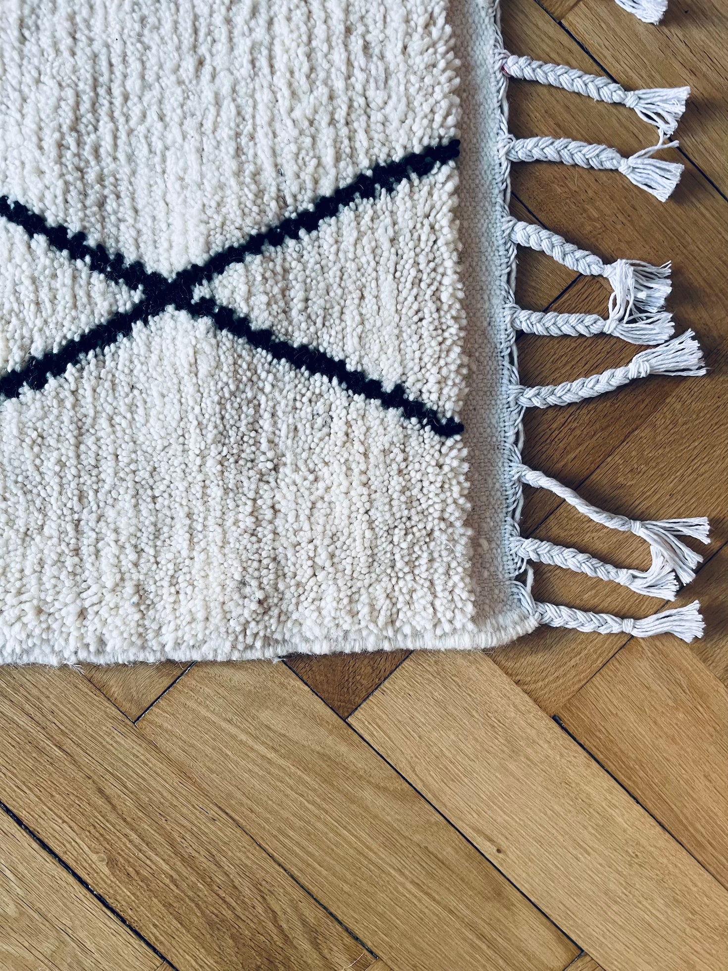 Eine Nahaufnahme des BERBER TEPPICH - MAHIMA (1,50 x 2,50m) von THE DANISH GIRL INTERIOR zeigt seine weiße Wolle mit schwarzen Linien und geflochtenen Quasten, handgeknüpft aus Naturwolle, auf hellem Holzparkett.