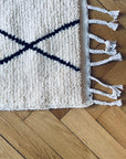 Eine Nahaufnahme des BERBER TEPPICH - MAHIMA (1,50 x 2,50m) von THE DANISH GIRL INTERIOR zeigt seine weiße Wolle mit schwarzen Linien und geflochtenen Quasten, handgeknüpft aus Naturwolle, auf hellem Holzparkett.