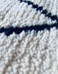 Nahaufnahme von THE DANISH GIRL INTERIOR BERBER TEPPICH - MAHIMA (1,50 x 2,50m), handgeknüpft aus natürlicher Wolle in Weiß mit schwarzen linearen Mustern. Oberflächendetails und Fasern sorgen für einen weichen, taktilen Look.