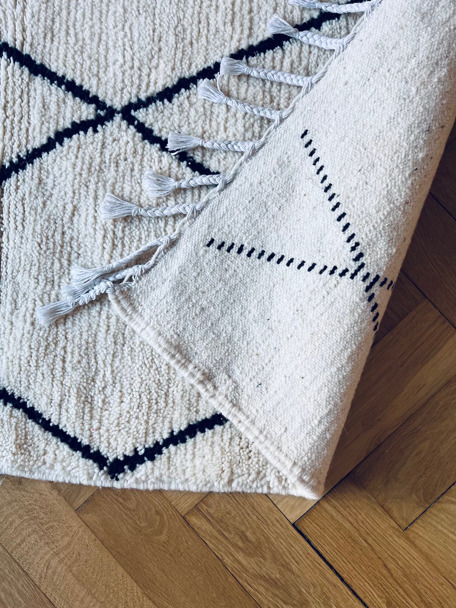 Eine Nahaufnahme des BERBER TEPPICH - MAHIMA (1,50 x 2,50m) von THE DANISH GIRL INTERIOR-geknüpft aus naturbelassener Wolle mit schwarzen Linien, Ecke gefaltet und Fransen sichtbar, auf Parkettboden.