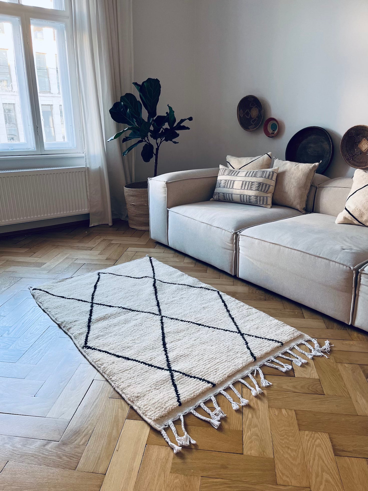 Ein helles Wohnzimmer mit einer beigen Couchgarnitur, gemusterten Kissen, einer Topfpflanze und Wanddekoration. Der BERBER TEPPICH - MAHIMA von THE DANISH GIRL INTERIOR (1,50 x 2,50m) mit schwarzen Rautenlinien liegt auf dem Parkett neben großen Fenstern mit durchsichtigen Vorhängen.