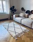 Ein helles Wohnzimmer mit einer beigen Couchgarnitur, gemusterten Kissen, einer Topfpflanze und Wanddekoration. Der BERBER TEPPICH - MAHIMA von THE DANISH GIRL INTERIOR (1,50 x 2,50m) mit schwarzen Rautenlinien liegt auf dem Parkett neben großen Fenstern mit durchsichtigen Vorhängen.